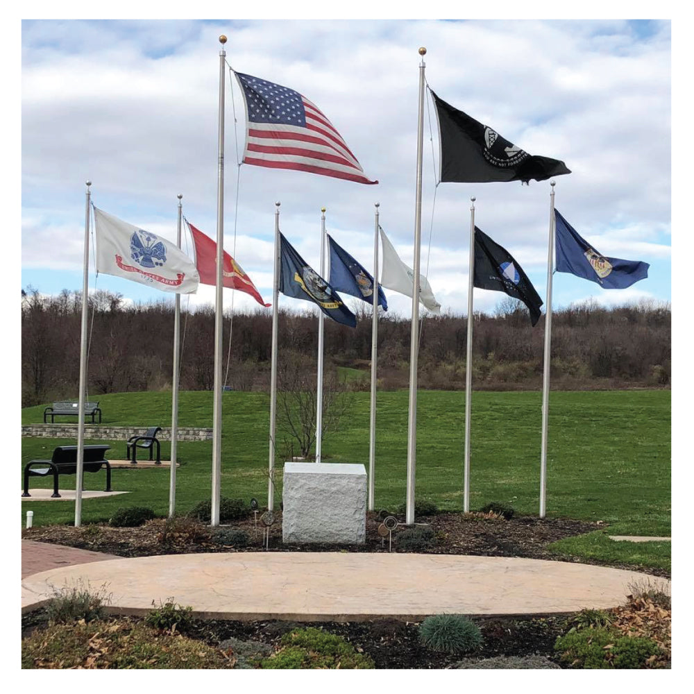 Multiple flags on flag poles waving in the wind in front of benches, grass and a hill of bare bushes