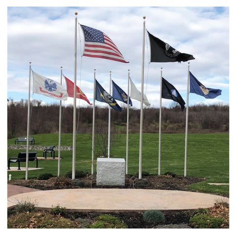 Multiple flags on flag poles waving in the wind in front of benches, grass, and a hill of bare bushes